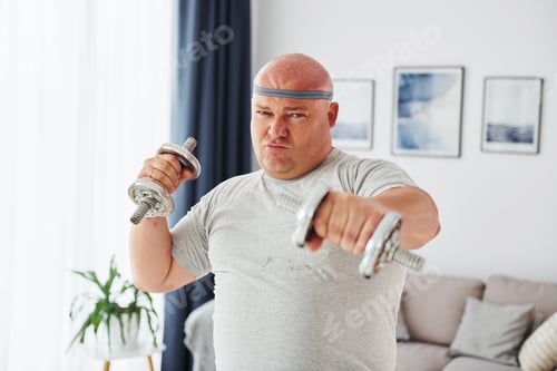 Preview: Uses dumbbells. Funny overweight man in casual clothes is indoors at home
