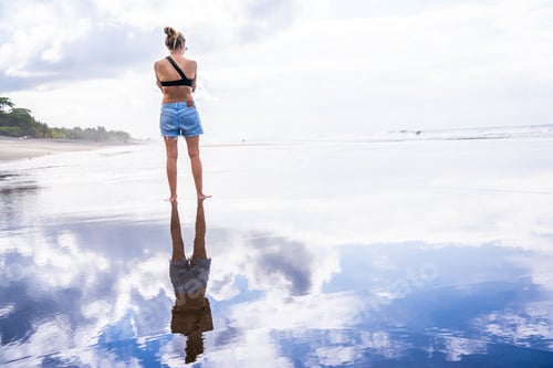 Preview: Woman on a beach while her silhouette and the sky are reflected in the water