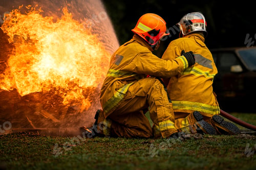 Preview: Two firefighters kneel in position while directing a powerful water stream at a large blaze