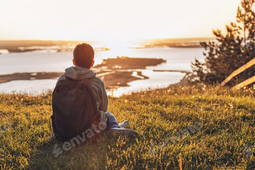 Preview: Rear view of tourist with backpack sitting on hill in grass field and enjoying beautiful sunset