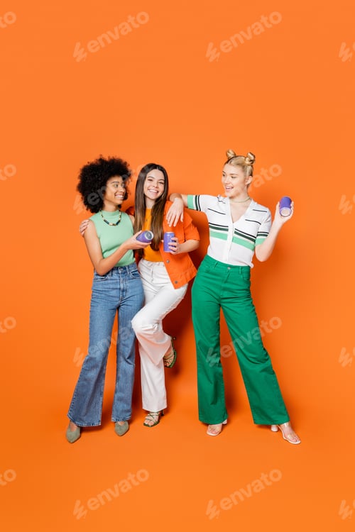 Preview: Full length of positive and multiethnic teenage girlfriends in stylish outfits holding canner drink
