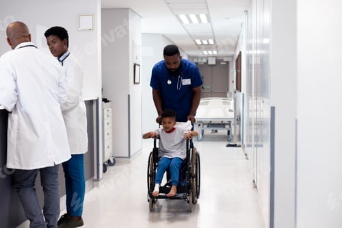 Preview: Vertical of african american male doctor pushing boy patient in wheelchair in corridor