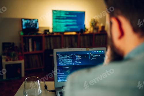 Preview: Rear view of an IT specialist sitting at home at night and working on software development.