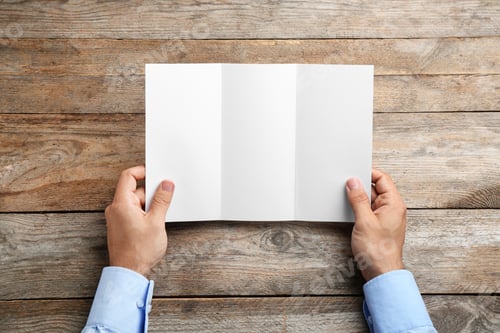 Preview: Young man holding blank brochure at wooden table, top view. Mock up for design