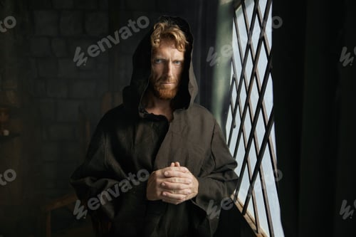 Preview: Pensive monk in medieval costume with hood looking through window of monastery