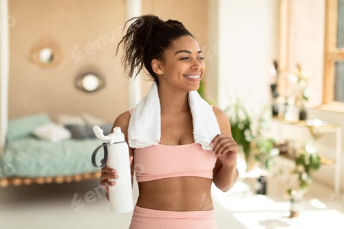 Preview: Smiling african american lady in sportswear having break while exercising at home, holding bottle