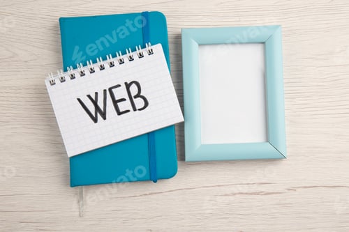 Preview: top view web written on spiral notebook on blue notepad empty photo frame on table