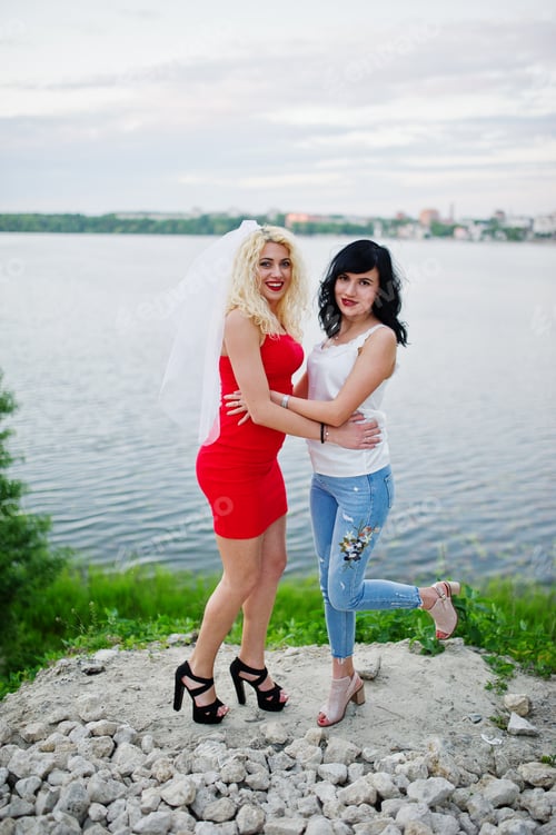 Preview: Awesome bride in red dress posing with her bridesmaid on the lakeside.