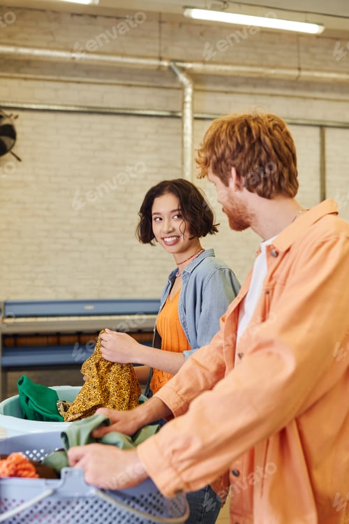 Preview: cheerful young asian woman talking to boyfriend near clothes and baskets in coin laundry