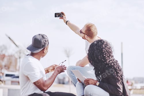 Preview: Beautiful multi ethnic friends using a laptop in the Street and make selphie