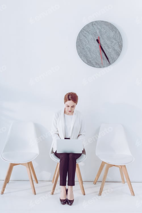 Preview: attractive woman using laptop while sitting on chair near clock