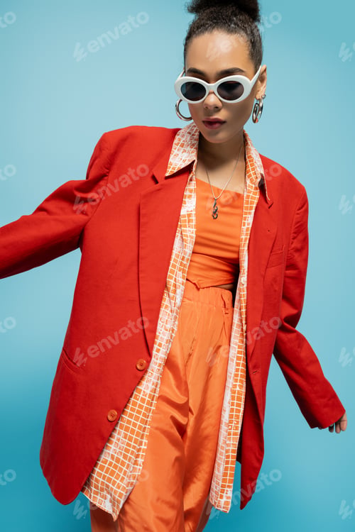 Preview: charismatic african american woman in trendy sunglasses and vibrant outfit posing on blue background