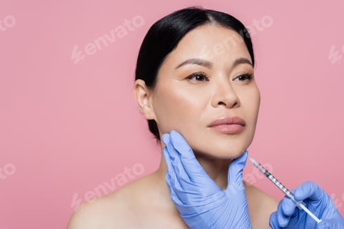 Preview: Cosmetologist in latex gloves holding syringe near chin of asian woman isolated on pink