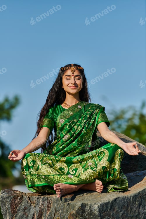 Preview: smiling indian barefoot woman in sari meditating on stone with blue sky on background