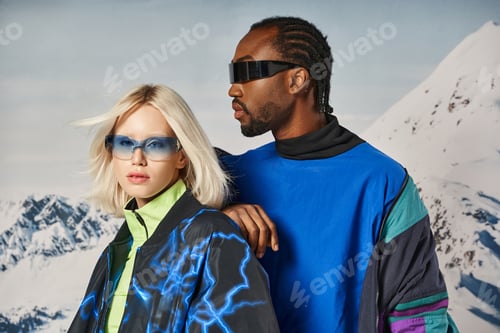 Preview: attractive couple in stylish sunglasses and warm outfits posing with snowy backdrop, winter concept