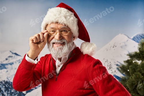 Preview: joyful man dressed as Santa in red hat touching his glasses and looking at camera, Merry Christmas