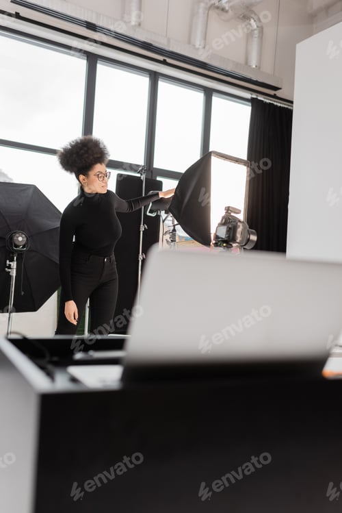 Preview: african american content maker in eyeglasses near softbox reflector in photo studio on blurred