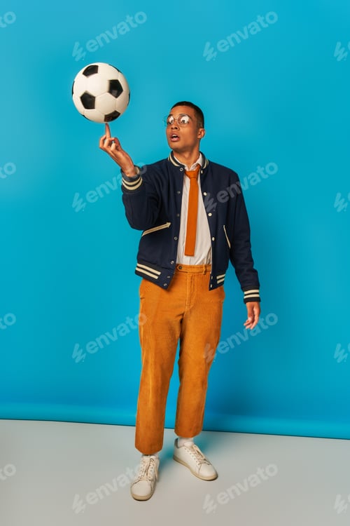 Preview: african american student in jacket and orange pants playing with soccer ball on blue