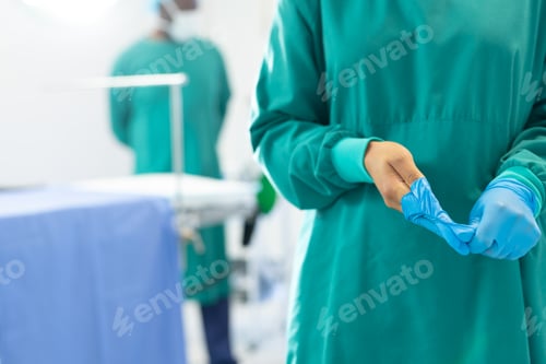 Preview: Midsection of female surgeon in surgical gown taking off gloves in operating theatre, copy space