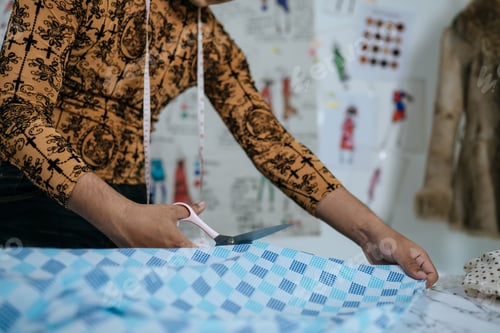 Preview: Cropped image of Asian young male tailor cutting piece of cloth at table in fashion studio.