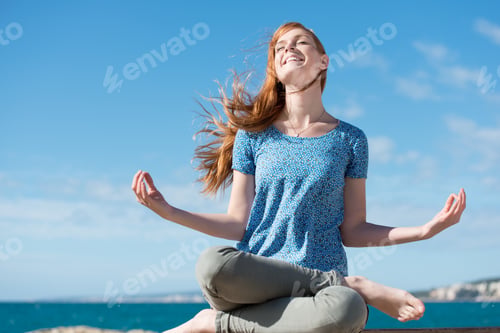Preview: Beautiful Woman Meditating At The Seaside Sitting Cross-Legged On The Rocks With Her Head Tilted To