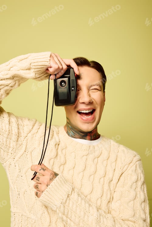 Preview: A joyful young man with tattoos playfully captures memories with a camera against a yellow backdrop