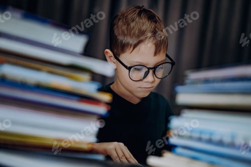 Preview: cute caucasian boy in eye glasses reading a book. Cheerful boy peeking from behind piles of books