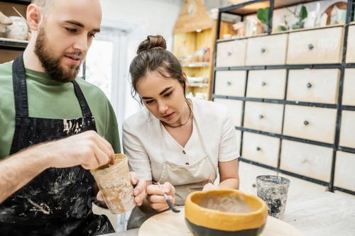 Preview: Craftsman in apron coloring clay bowl with blurred girlfriend together in ceramic workshop