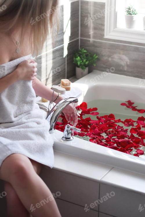 Preview: Young woman is checking water while running a bath in a bathtub filled with red rose petals