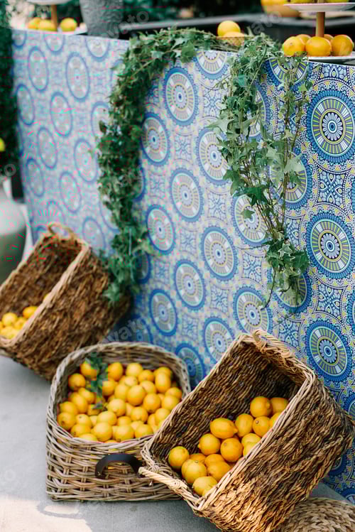 Preview: Wicker baskets with tangerines and lemons stand near a table covered with a colorful tablecloth and