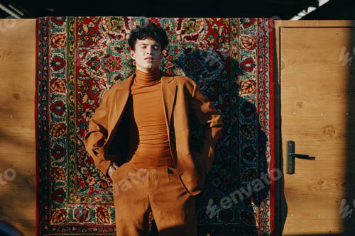 Preview: Young man in a brown suit stands confidently against an ornate patterned background His expression