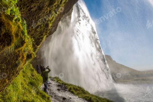 Preview: People standing on a narrow path underneath a waterfall cascade over a sheer cliff.