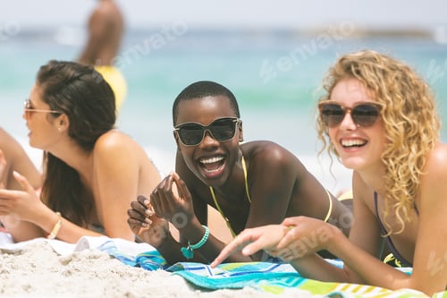 Preview: Front view of multi ethnic women interacting with each other while lying at beach on sunny day