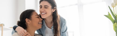 Preview: cheerful lesbian woman with closed eyes hugging multiracial girlfriend at home, banner