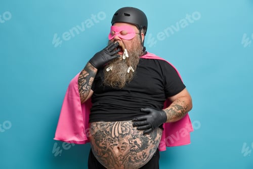 Preview: Exhausted male superhero yawns and covers mouth stands with big tattooed belly indoor being tired of