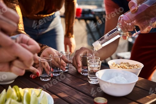 Visualização: cropped view of multicultural friends in casual attires pouring tequila shots while at rooftop party