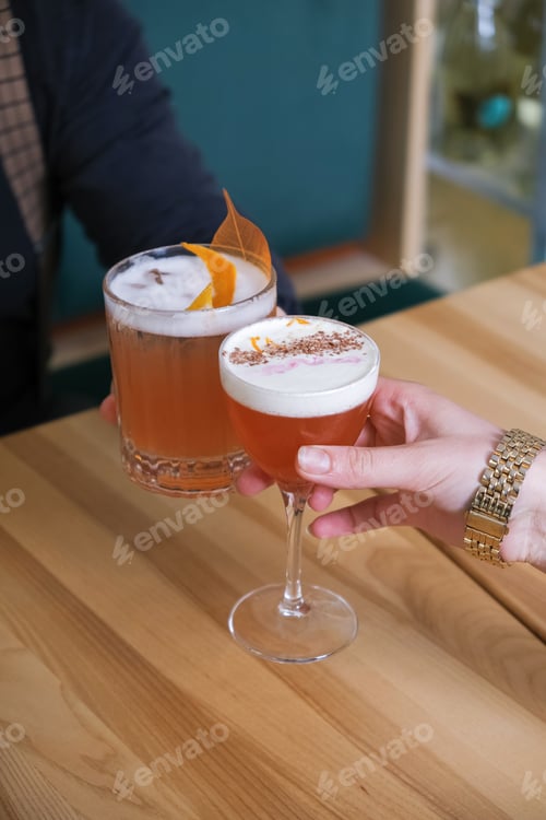 Visualização: Dois coquetéis alcoólicos nas mãos sobre uma mesa de cafeteria de madeira.