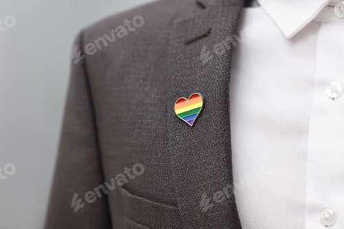 Preview: Man with heart shaped pin in colors of LGBT flag indoors, closeup