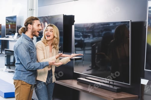 Preview: shocked boyfriend and girlfriend pointing with hands at tv in home appliance store