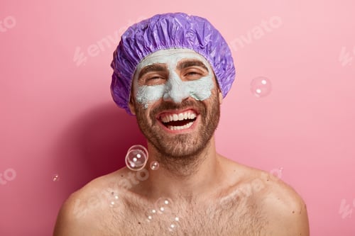 Preview: Photo of satisfied European man enjoys taking shower, wears bathcap, laughs happily, applies facial