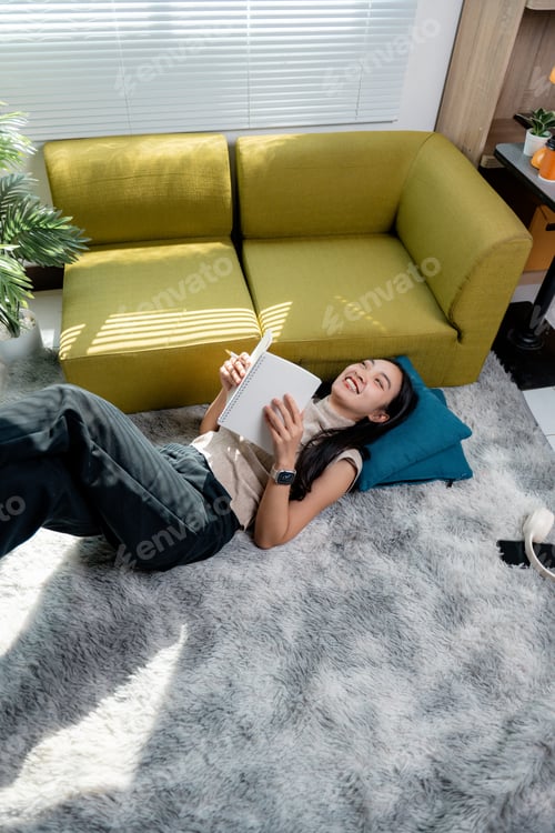 Preview: Freelancer woman relaxing on carpet, writing notes in notebook during leisurely work moment
