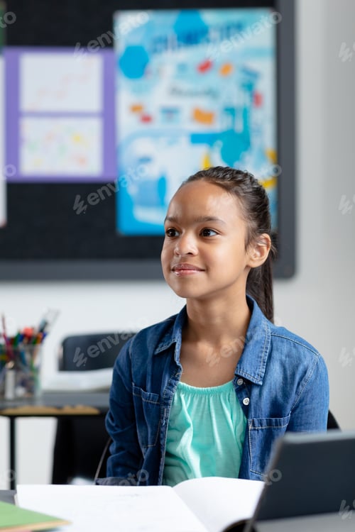 Preview: Vertical of happy biracial schoolgirl sitting at desk in class, listening, with copy space