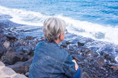 Preview: Rear view of relaxed caucasian senior woman sitting on the beach looking at the horizon