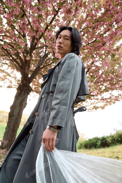 Preview: Stylish young man in grey trench coat strolls through park adorned with cherry blossoms