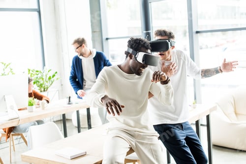Preview: two cheerful multicultural colleagues gesturing while using vr headsets in office