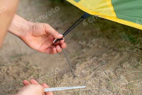 Preview: person is kneeling on the ground, securing a tent peg to the ground near a tent.