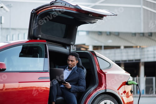 Preview: African man working on laptop while electric car is charging