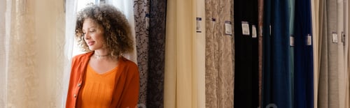 Preview: young woman with curly hair standing near variety of curtains in textile shop, banner
