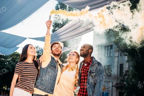 Preview: interracial cheerful young friends with colorful smoke bomb in park