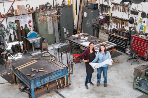 Preview: High angle view of two women standing in metal workshop, holding technical blueprint.